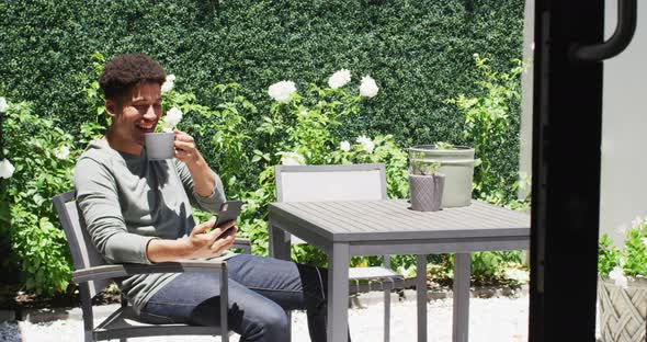 Happy biracial man using smartphone and drinking coffee in garden