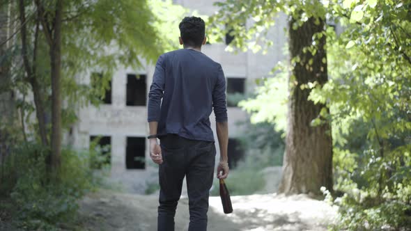 Back View of Frustrated Young Mixed-race Man Walking Along Slum with Alcohol Bottle. Camera Follows