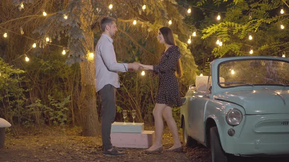 Wide Shot of Cheerful Caucasian Boyfriend and Girlfriend Dancing on Summer Evening with Garland