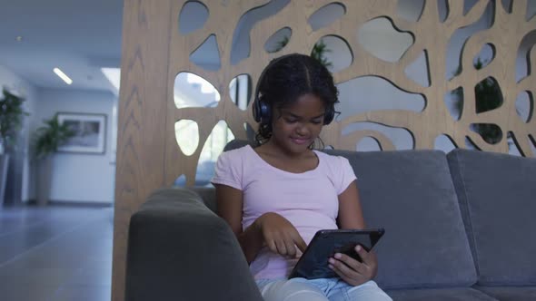 African american girl wearing headphones using digital tablet while sitting on the couch at hospital