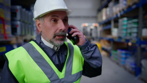 Confident Professional Senior Warehouse Manager in Hard Hat Talking on the Phone As Blurred Loader