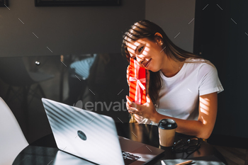 young woman opening gift in front of laptop during video call or chat, celebrating birthday online