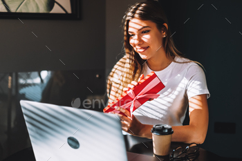 young woman opening gift in front of laptop during video call or chat, celebrating birthday online