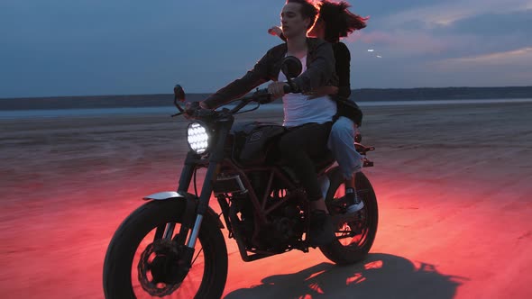 Young Couple Riding on Vintage Motorcycle with Red Burning Signal Fire After Sunset on Beach Slow