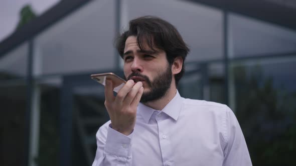 Concentrated Handsome Bearded Man Recording Voice Mail on Smartphone Standing on Office Terrace
