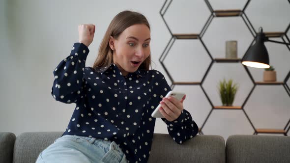 Happy Young Woman Looking at the Smartphone Screen Celebrating Winning an Online Bet Sitting at Home