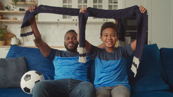 Cheerful Black Father and Son with Football Scarves Watching Soccer Game on Tv