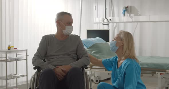 Female Nurse Wearing Surgical Mask Talking to Male Patient Sitting in Wheelchair in Hospital Ward