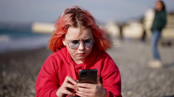 Little Girl is Resting on Seashore in City and Playing Video Game in Smartphone