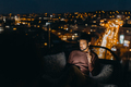 Young African AMerican man sitting on balcony with urban view and using tablet at night - PhotoDune Item for Sale