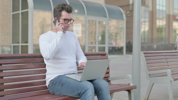Angry Young Man Talking on Phone and Using Laptop While Sitting Outdoor on Bench