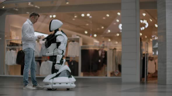 A Man in a Shirt Communicates with a White Robot Asking Questions and Pressing the Screen with His