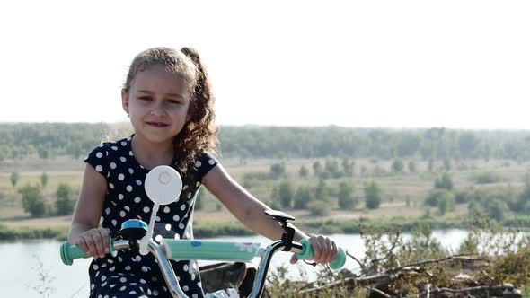 Cute Little Happy Girl Sitting on a New Bike on the Top of the Hill, Looking at the Camera and