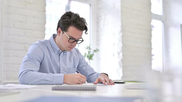 Serious Working Young Man Writing on Paper in Modern Office