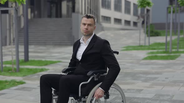 Serious Businessman in Classical Suit Sitting in Wheel Chair and Riding at City Street