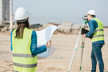 Selective focus of surveyor with blueprint and colleague using digital level on construction site