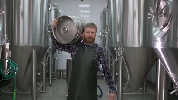 Worker Male Brewer Carries a Keg Filled with Beer Passing Beer Tanks