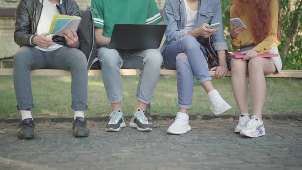 Legs of Four Caucasian University Students Sitting on Bench Outdoors Surfing Internet and Doing