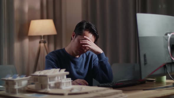 Asian Male Engineer With The House Model Having A Headache While Working On A Desktop At Home