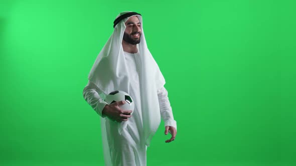 Cheerful Arab Man in a White Robe on the Green Background Wearing Kandura and Walks Holding a Soccer