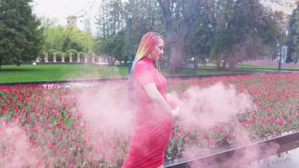 A Girl with Makeup with Rainbow Braids in Red Dress Posing in Red Smoke Against the Background of a