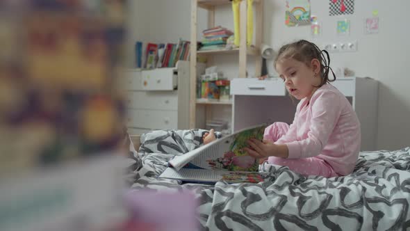 Little Baby Girl Reading a Book on the Bed in the Nursery