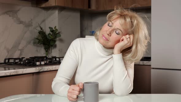 Sleepy Middle Aged Woman Drinks Coffee Sitting in Kitchen