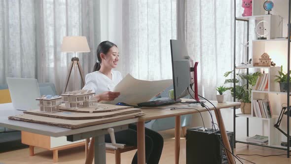 Woman Engineer With The House Model Opening And Looking At The Blueprint While Working On A Desktop