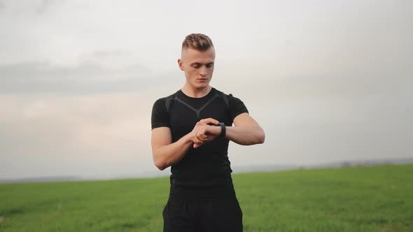 The Athlete is Standing on the Field and Choosing the Type of Training on His Watch