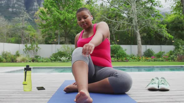 Happy african american plus size woman practicing yoga, stretching next to swimming pool in garden