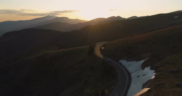 Drone Follows Lonely Car Driving on Majestic Winding Mountain Road at Golden Sunset