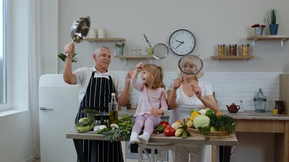 Senior Woman and Man with Grandchild Girl Making a Funny Dance with Strainer and Vegetables at Home