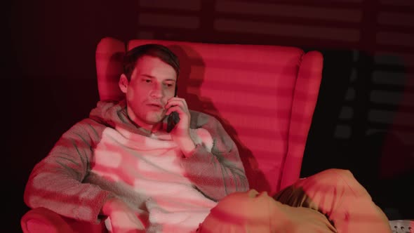Young Man Speaks on Smartphone Sitting in Armchair in Dark Room with Falling Red Light and Shadow