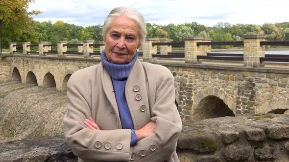 An Elderly Woman Leans Against Stone Barrier, Crosses Her Arms Across Her Chest and Looks Seriously