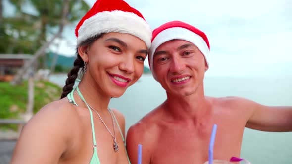 Happy Couple Smiling at Camera Taking Self-portrait Wearing Santa Hats on Beach Vacation