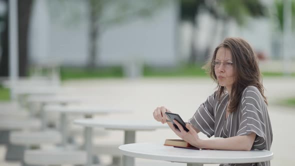 Serious Woman with Glasses is Reading Book and Viewing Learning Video in Smartphone Outdoors