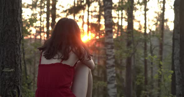 a Girl in a Red Dress Sits in the Woods and Looks at the Sunset View From the Back
