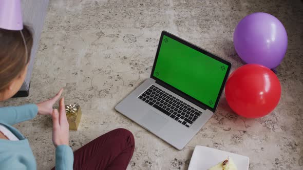 Surprised woman in party hat holding a gift box while having a video call on laptop at home