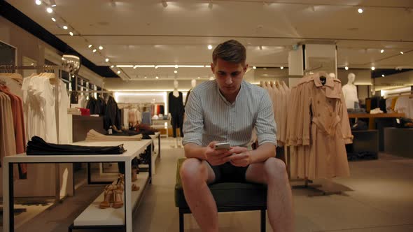 Caucasian Man Leafing Through Social Networks in the Phone in a Clothing Store