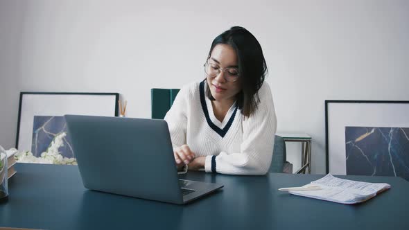 Young Asian Woman Employee in Glasses is Typing on Laptop and Smiling While Working at Office