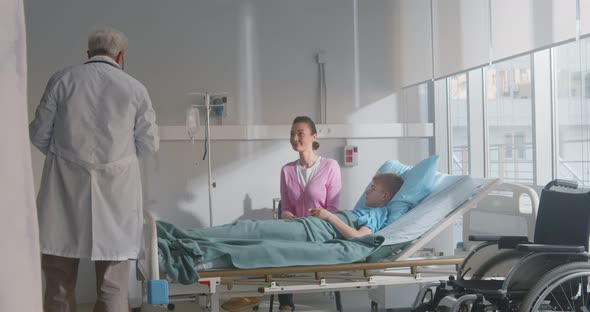 Mature Doctor Talking to Young Mother and Sick Son Lying in Hospital Bed