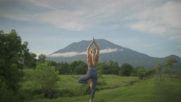 Athletic blond woman in yoga tree pose balancing on rock, Mount Agung in background