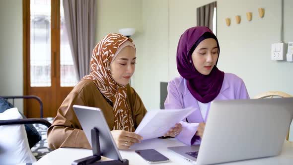 Two asian muslim women working together with using laptop pc tablet and paper chart in living room