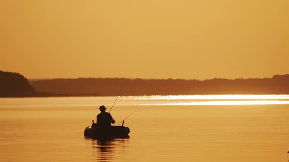 Silhouette of a man in hat fishing from the boat on the evening river background.