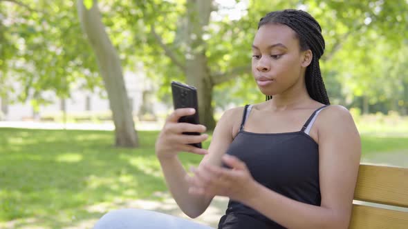 A Young Black Woman Takes Selfies with a Smartphone and Sits on a Bench in a Park on a Sunny Day