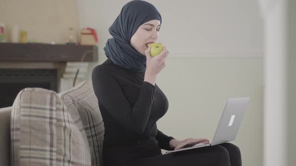 Young Muslim Woman Working or Chatting By Laptop and Biting Juicy Apple Wearing Traditional