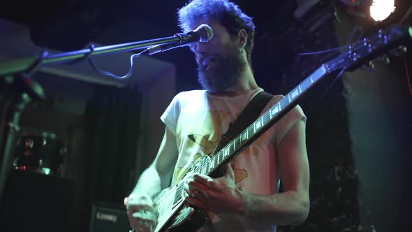 From Below View of Modern Bearded Man in T-shirt Singing in Mic and Playing Electric Guitar in Light