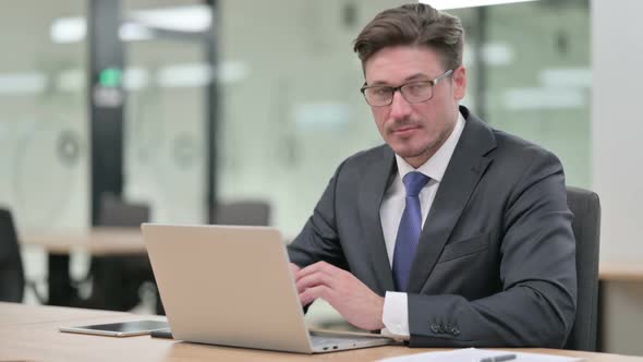 Middle Aged Businessman with Laptop Smiling at Camera in Office