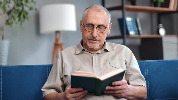 Happy Mature Old 70s Man Gray Haired Man Reading Interesting Paper Book Resting on Couch Retirement