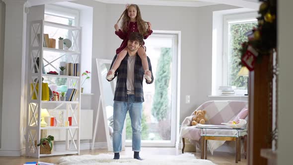 Wide Shot Portrait of Happy Young Father Holding Cheerful Cute Daughter on Shoulders Standing in